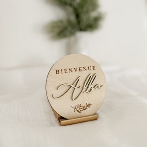 May include: A round wooden sign with the words "BIENVENUE Alba" engraved on it. The sign is light brown and has a small floral design. It is displayed on a small wooden stand.