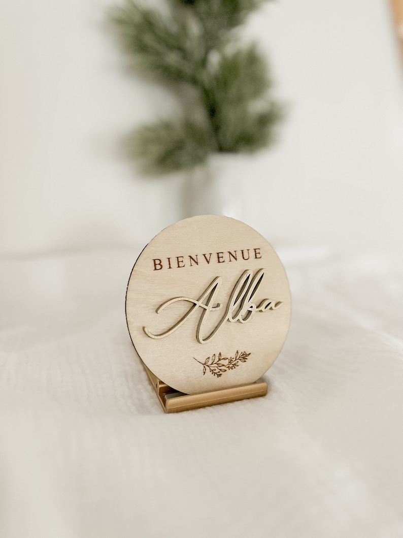 May include: A round wooden sign with the words "BIENVENUE Alba" engraved on it. The sign is light brown and has a small floral design. It is displayed on a small wooden stand.