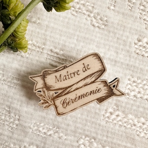 Peut inclure: Une épingle en bois avec l'inscription "Maître de Cérémonie" dans un motif de bannière. L'épingle est marron clair avec du texte marron foncé et un petit détail de feuille. L'épingle est sur une surface blanche texturée.