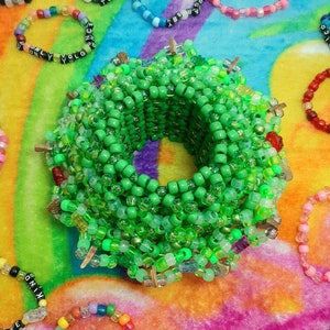 Puede incluir: Un brazalete de cuentas verdes con un diseño de cruz. El brazalete está hecho de muchas capas de cuentas y está rodeado de otros brazaletes de cuentas coloridos con palabras en ellos.