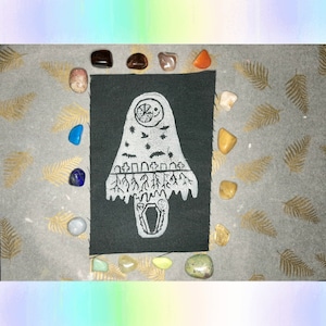 May include: Black fabric patch featuring a white design of a ghost-like figure with a moon and stars inside. The ghost is standing over a graveyard with trees and a coffin.