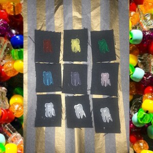 May include: Nine fabric patches with a tooth design in different colors. The patches are arranged in a 3x3 grid on a striped background. The colors of the teeth are red, yellow, green, blue, purple, pink, white, gray, and black.