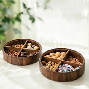 May include: Two circular wooden serving platters, each with four sections, filled with an assortment of nuts and sweets. The dark brown wood trays are on a white surface. The background features foliage and natural light.