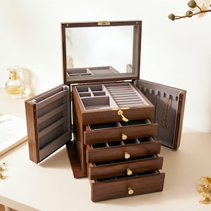 Puede incluir: Una caja de joyería de madera marrón oscuro con tapa espejada y múltiples cajones. La caja está abierta, revelando compartimentos y cajones forrados de fieltro gris. Los herrajes dorados acentúan la caja, diseñada para guardar joyas.