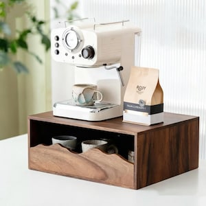Könnte beinhalten: Eine cremefarbene Espressomaschine steht auf einer rechteckigen Aufbewahrungsbox aus dunklem Holz mit gewellter Vorderkante. Eine Keramiktasse steht auf der Abtropfschale der Maschine, daneben eine Packung Roby Italian Coffee Blend. In der Box befinden sich zwei Tassen.