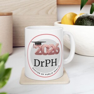 May include: White ceramic mug featuring a graduation cap and the text "2026 DrPH Doctor of Public Health" in a circular design. The mug is on a beige coaster, with a blurred background.