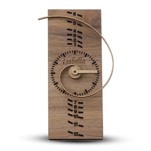 May include: A wooden clock with a spiral design and the name "Isabella" on the face. The clock has a wooden hand and a wooden frame.