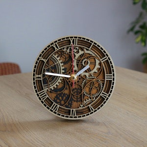 Puede incluir: Un reloj redondo de madera con números romanos y un diseño de engranajes intrincados. El reloj tiene una aguja de segundos roja, agujas de horas y minutos blancas y un centro dorado.