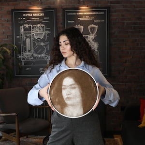 Pode incluir: Uma mulher segura um retrato redondo em tons sépia com uma moldura branca. O retrato retrata o rosto de uma mulher. O fundo apresenta paredes de tijolo, diagramas emoldurados e uma área de estar.