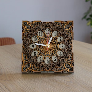 Puede incluir: Un reloj cuadrado de madera con un intrincado diseño de corte por láser. La esfera del reloj presenta un patrón de mandala con números romanos y manecillas negras.