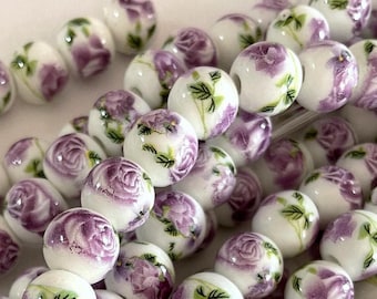 Può includere: Una collana di perline in ceramica bianca con un motivo floreale viola. Le perline sono rotonde e hanno una finitura lucida.