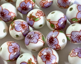 Può includere: Una collezione di perline in ceramica bianca con disegni floreali viola e rosa. Le perline sono rotonde e hanno una finitura lucida.
