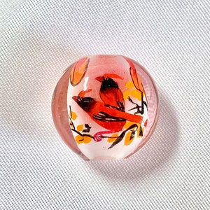 May include: A clear glass bead with a painted image of a red cardinal bird perched on a branch with yellow leaves. The bead has a pink tint.