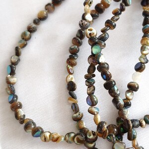 May include: A strand of irregular, small beads crafted from natural abalone shell. The beads feature a dark brown base color with iridescent flashes of blue, green, and pearlescent white. The strand is arranged in a loose, overlapping circular pattern.