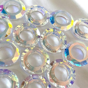 Puede incluir: Una colección de anillos de cristal transparente iridiscente con un agujero en el centro. Los anillos son de varios tamaños y están dispuestos en un semicírculo sobre una superficie blanca.