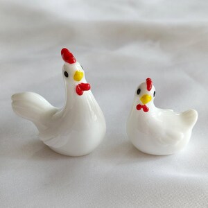 Peut inclure: Deux figurines de poulet en verre blanc, l'une plus grande que l'autre. Les deux poulets présentent des corps blancs lisses, de petits yeux noirs, des becs jaune vif, ainsi que des crêtes et des barbillons rouge éclatant en verre brillant.