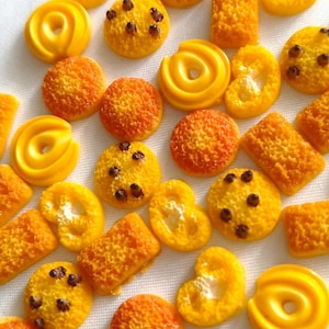 May include: A collection of miniature yellow and orange food-shaped cabochons, including round cookies with dark brown chocolate chips, smooth yellow ring-shaped pastries, textured rectangular biscuits, and irregular yellow pastry shapes.