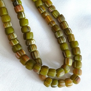 Puede incluir: Un collar de cuentas de vidrio verde oliva con patrones de rayas verticales blancas y naranjas. Las cuentas son cilíndricas con bordes redondeados y están dispuestas en una formación curva, mostrando una paleta de colores uniforme.