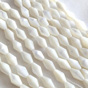 Puede incluir: Múltiples hilos de cuentas de nácar blanco dispuestas en líneas paralelas. Cada cuenta presenta una forma de diamante facetado con un acabado liso e iridiscente y un orificio central para ensartar, creando una apariencia uniforme y brillante.