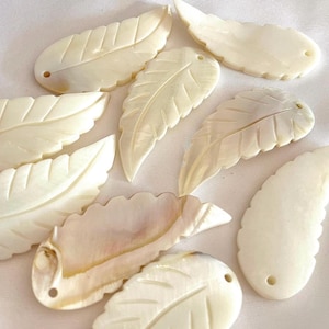 Puede incluir: Una colección de diez colgantes de nácar blanco en forma de hoja con un agujero en la parte superior para encordar. Las hojas están talladas con un patrón de vetas detallado.