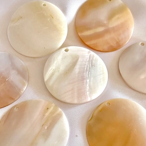 May include: A collection of eight round, iridescent, mother-of-pearl shell discs with a hole in the center. The discs are various shades of white, cream, and beige.
