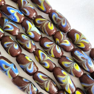 Puede incluir: Un hilo de cuentas cilíndricas de vidrio con un color base marrón oscuro y un patrón jaspeado y arremolinado de acentos en amarillo brillante y azul claro. Las cuentas están dispuestas en filas paralelas, mostrando un acabado liso y brillante.