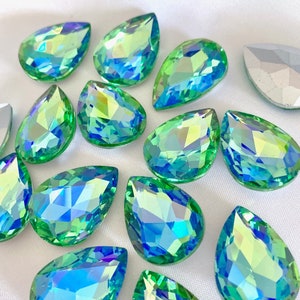 Op de afbeelding: Een verzameling van druppelvormige, iriserende glazen strasssteentjes in tinten groen en blauw. De strasssteentjes hebben een facetgeslepen ontwerp en zijn verspreid over een wit oppervlak.