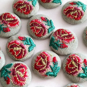 以下が含まれることがあります： 刺繍された花柄が特徴の布張り丸ボタンのコレクション。各ボタンはライトグレーのテクスチャーベースで、鮮やかな赤とピンクのバラのモチーフが施され、緑の葉と金色の糸のディテールが全体にアクセントとしてあしらわれています。