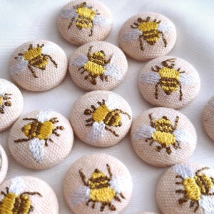 以下が含まれることがあります： 刺繍されたハチのデザインが施された、布で覆われた丸いボタンのコレクション。各ボタンはライトベージュのベースで、黄色と茶色のハチのモチーフと白い羽が特徴です。1つのボタンは裏返されており、平らな銀色の金属製の裏面が見えています。