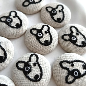 May include: A collection of round, fabric-covered buttons featuring a black embroidered outline of a dog face. Each button is made of a light beige, textured linen-like material, showcasing a consistent graphic design of a canine head with one eye.