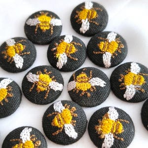 以下が含まれることがあります： 黒い布地をベースに、黄色と白の刺繍で蜂のデザインが施された円形のボタンのコレクション。各ボタンには、黄色い体と白い羽を持つ蜂のモチーフが詳細に描かれており、白い表面に散らばるように配置されています。