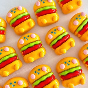 Könnte beinhalten: Eine Sammlung von 11 gelben und grünen Miniatur-Hamburger-Anhängern mit roten und weißen Akzenten. Die Anhänger sind aus Polymer Clay gefertigt und haben einen niedlichen Cartoon-Stil.