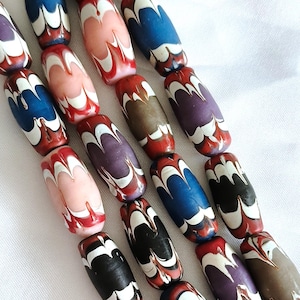 May include: A collection of cylindrical glass beads arranged in three vertical strands. Each bead features a distinct chevron pattern with alternating bands of white, red, and solid colours including blue, purple, pink, black, and brown.