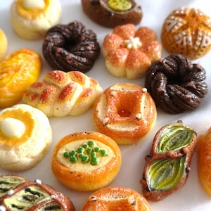 Puede incluir: Una colección de dijes de comida en miniatura con forma de diversos productos horneados, incluyendo bollos redondos, pasteles trenzados, donas de chocolate oscuro y panes ovalados con coberturas verdes o amarillas, con texturas realistas y acabados brillantes.