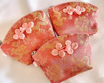 May include: Three small pink fabric pouches with gold embroidery and pink fabric bows. The pouches have a zipper closure.
