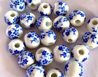 Puede incluir: Una colección de cuentas de cerámica blancas y redondas con un patrón floral azul pintado a mano. Cada cuenta tiene un orificio central para enhebrar. La superficie de las cuentas es lisa y brillante, mostrando un esquema de color azul y blanco.