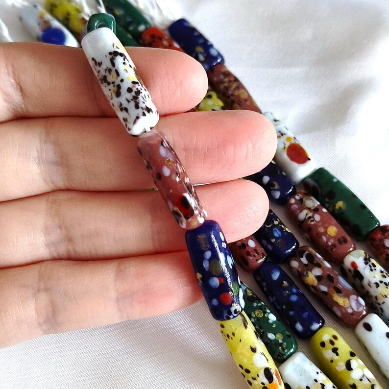 May include: A collection of cylindrical glass beads arranged in multiple strands. Each bead features a unique speckled or mottled pattern in various colours including white, blue, red, yellow, green, and brown, with a smooth, glossy finish.