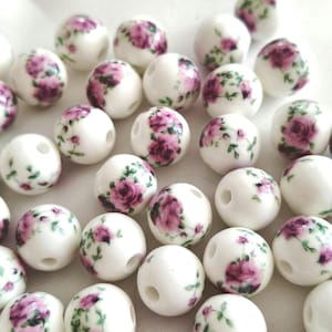Könnte beinhalten: Weiße Keramikperlen mit einem Blumendesign mit rosa Rosen und grünen Blättern. Die Perlen sind rund mit einem Loch in der Mitte, geeignet für die Schmuckherstellung oder Bastelarbeiten. Die Perlen sind verstreut.