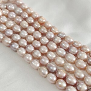 May include: Several strands of oval pearls in a palette of pink, lavender, and cream. The pearls are arranged in parallel rows, displaying their natural iridescence. The background is a soft, white fabric, accentuating the pearls' colours.