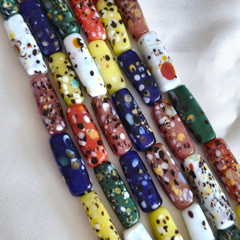 May include: A collection of cylindrical glass beads arranged in parallel rows. Each bead features a unique speckled pattern with various colours including white, blue, yellow, green, orange, and brown, often accented with small metallic gold flecks.
