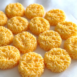 May include: A collection of small, round, yellow wax melts, each featuring a detailed, raised, noodle-like texture on the surface. These individual pieces are arranged in a group, showcasing a consistent golden-yellow color and a uniform, circular shape with intricate, swirling, pasta-inspired patterns across the top of each piece.