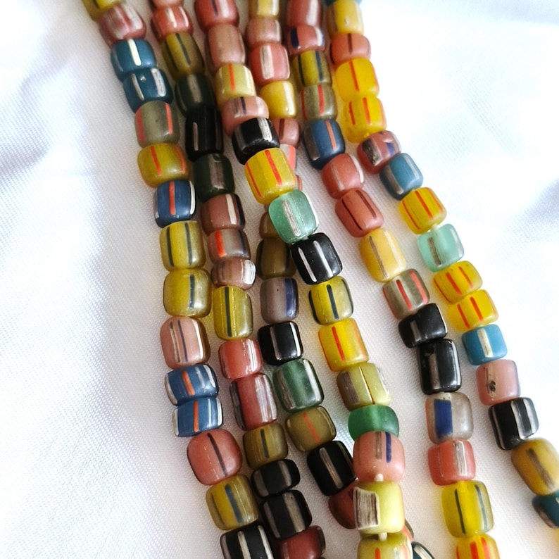 May include: Multiple strands of cylindrical glass beads in various colours including yellow, blue, black, green, and terracotta. Each bead features a distinct vertical stripe pattern in a contrasting colour, creating a textured and repetitive visual effect.