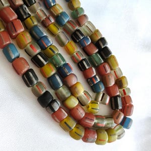 May include: A collection of multi-coloured glass beads arranged in several strands. The beads are square-shaped with rounded edges and feature a distinct vertical stripe pattern in contrasting colours including red, yellow, blue, black, and green.