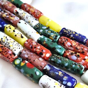 May include: A collection of cylindrical glass beads arranged in parallel rows. Each bead features a unique speckled or mottled pattern in various colours including red, green, blue, yellow, white, and brown, with a smooth, glossy finish.