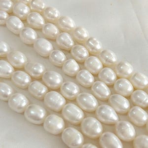 Puede incluir: Primer plano de varios hilos de perlas ovaladas, lustrosas y blanquecinas. Las perlas son uniformes en tamaño y forma, con una superficie lisa y pulida. El fondo es una tela blanca suave, que resalta el brillo natural de las perlas.