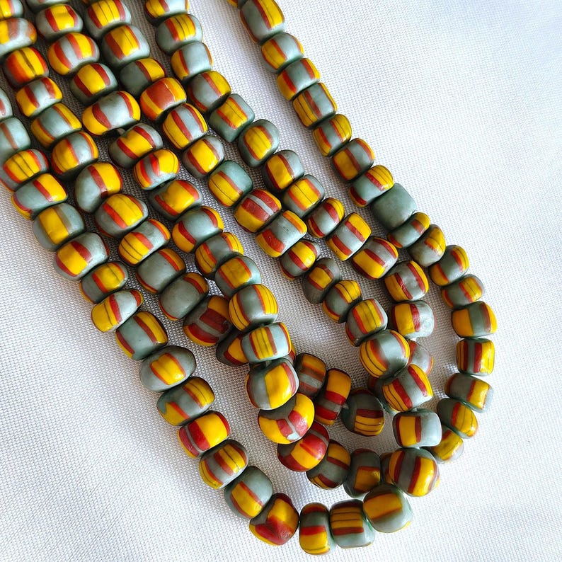 May include: Multiple strands of cylindrical glass beads featuring a matte sage green base colour with horizontal stripes in vibrant yellow and deep red. The beads are arranged in a series of parallel lines, showcasing a consistent pattern.