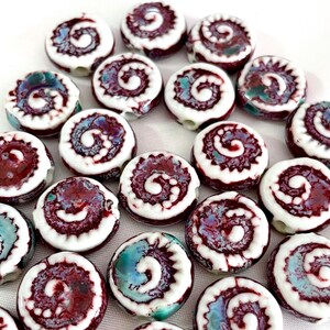 May include: A collection of ceramic beads with a spiral design. The beads are white with a dark red spiral pattern and accents of teal and purple. These beads are suitable for jewelry making and crafts.