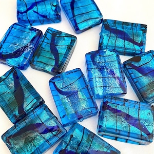 May include: A collection of rectangular, dichroic glass beads in shades of blue and teal. Each bead features a unique pattern of dark blue and black lines and swirls, creating a textured, iridescent effect. The beads are scattered on a white surface.
