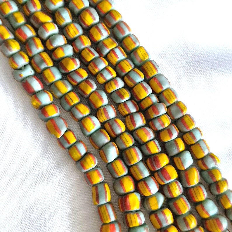 May include: A strand of small, square-shaped beads arranged in multiple vertical rows. Each bead features a light mint green base colour with a central vertical stripe of bright yellow and a thin, dark orange line running through the centre.