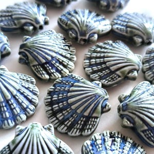May include: A collection of ceramic beads shaped like scallop shells. Each bead features a glossy finish with a color palette of deep blue, sage green, and off-white. The surface of each shell bead displays raised ridges and a textured pattern.